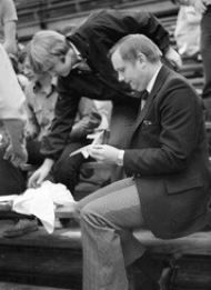 Neil Armstrong, wearing a suit, sits on a bleacher to create a paper airplane.
