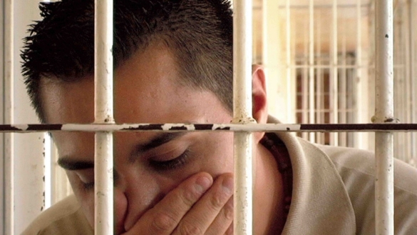 Man behind bars looks ay the ground and holds his hand over his mouth and nose