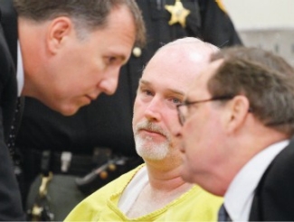 Mark Godsey and Jim Petro lean in to discuss Gillispie's case in front of him, wearing the yellow uniform of a prisoner.