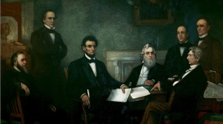 Lincoln is seated at a table with five other gentlemen, all in suits and ties, standing or sitting around him.