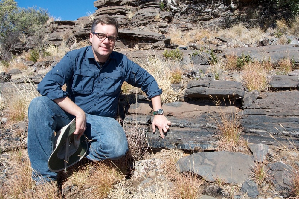 UC geologist, Andrew Czaja kneels beside a layer of ancient rock that contain fossilized bacteria.