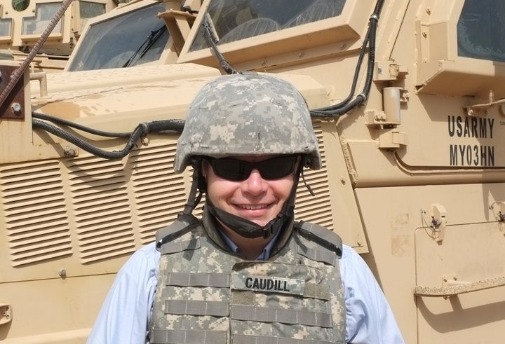 David Caudill wears bullet-proof vest and helmet, sanding in front of armored personnel carrier