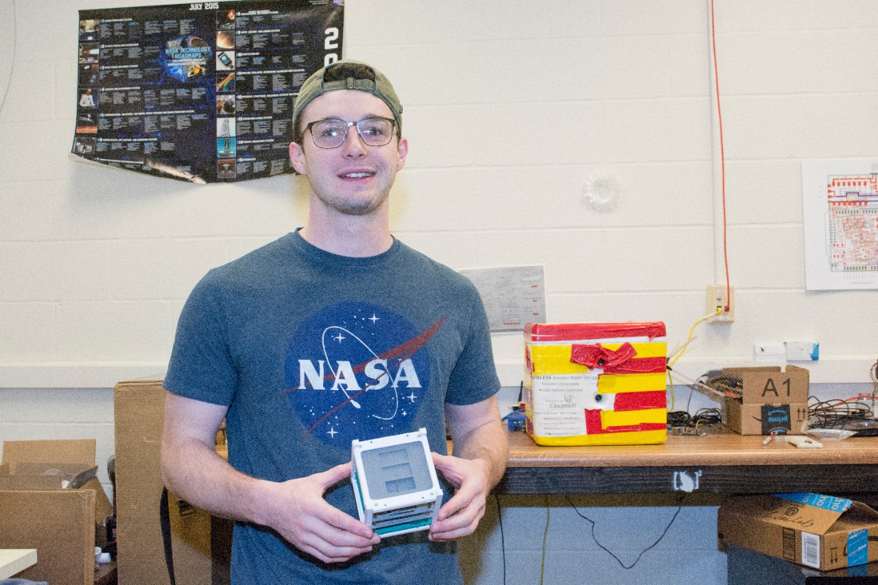 UC CubeCats founder Adam Herrmann holds a model of a cubesat in the club's lab. On the bench behind him sits the payload from the club's successful 2017 balloon launch.