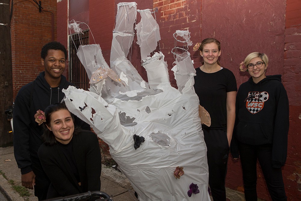 DAAP students display illuminated Body Mantle projects made from inexpensive materials and light-up components. Here, students in black pose with a giant white hand sculpture.