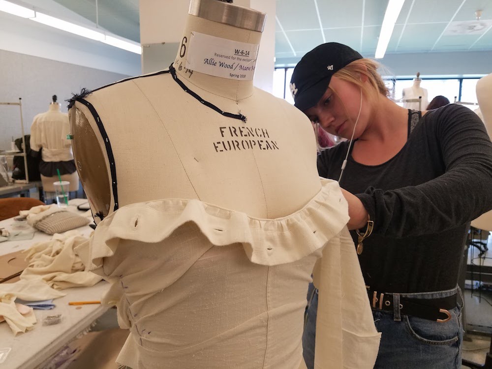 A student fits a garment made from two white button-up shirts on a dress form.