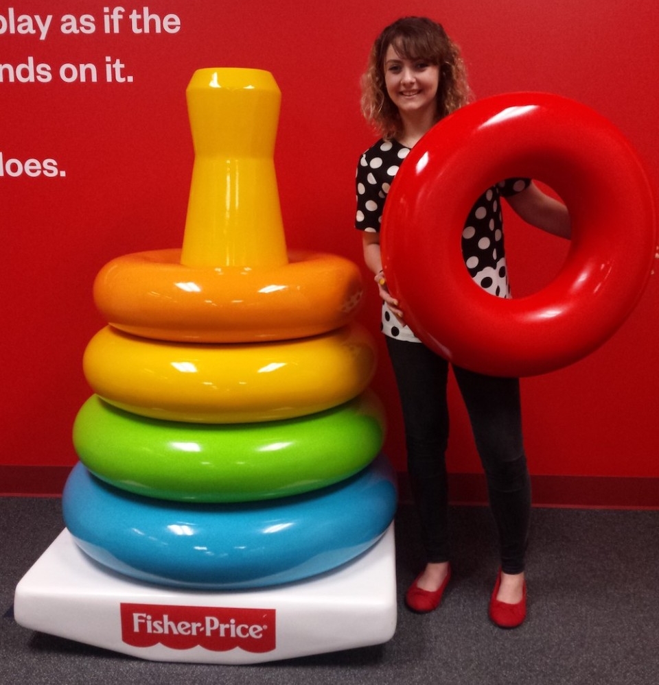 A young woman stands next to jumbo-sized Fisher-Price rainbow stacking rings toy.