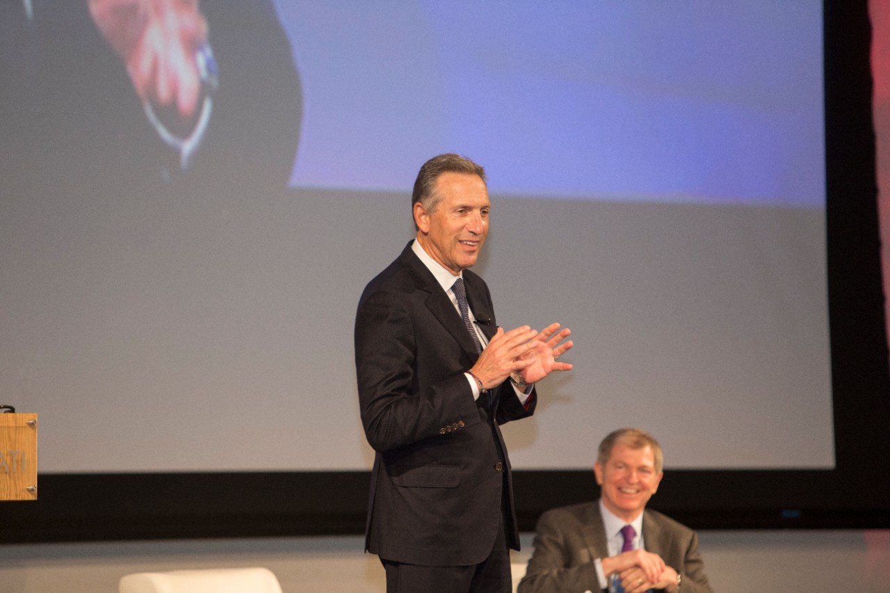 Starbucks Executive Chairman Howard Schultz stands on stage, with former J.C. Penney CEO Myron Ullman III looking on in the background, seated.
