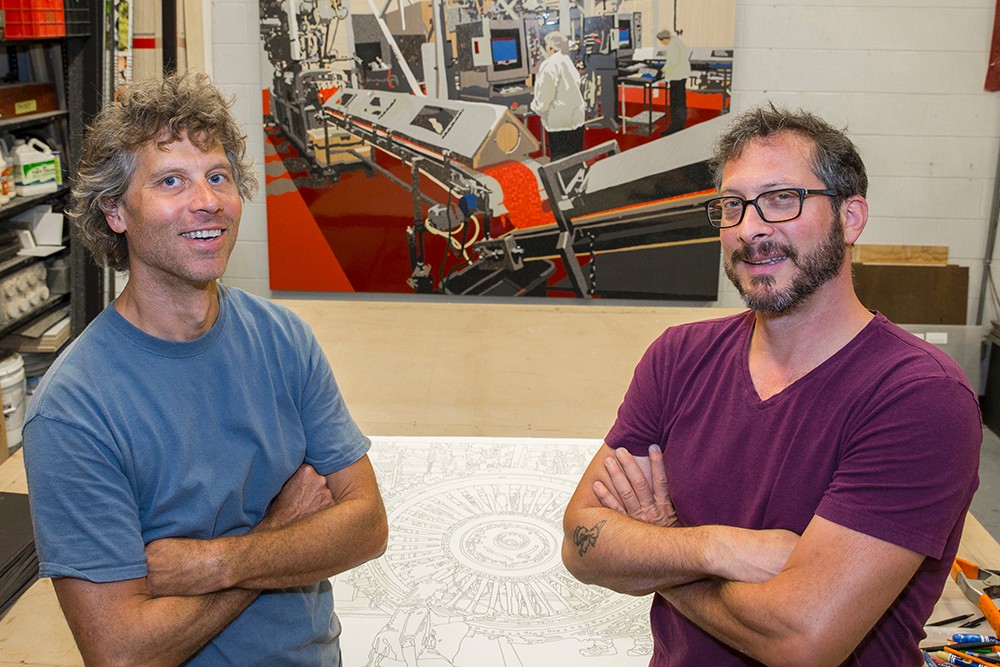 Matt Lynch and Curtis Goldstein pose in front of a line drawing of an engine from G.E. Aviation and a completed mural of the Airheads candy factory.