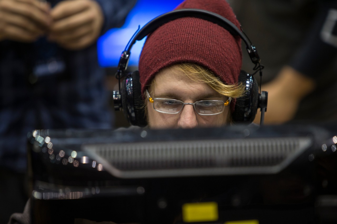 A bespectacled male student wearing a hat looks at a computer monitor.