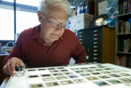 A older white gentleman with white hair leans very close into a light board scattered with photographic slides.