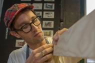 A young man in glasses and a plaid fabric cap works the grain of a fine piece of wood.