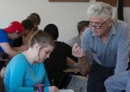 A University of Cincinnati professor, Ricardo Moena, with glasses at the end of his nose, leans in to a student in a classroom.