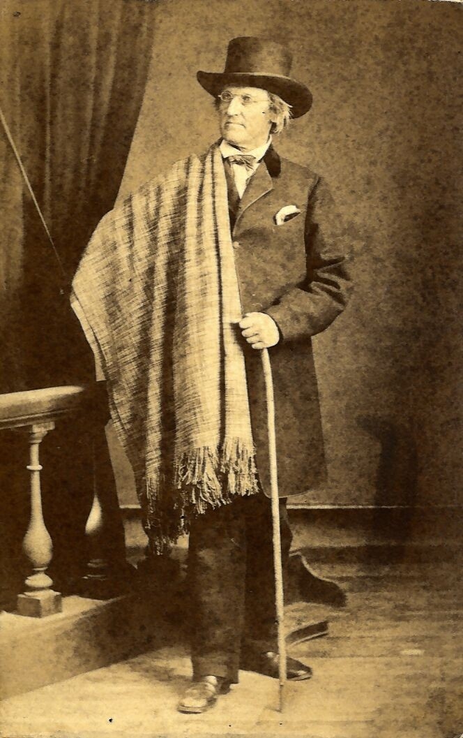 In a black and white photograph, John Hough James stands with a scarf, top hat and cane.