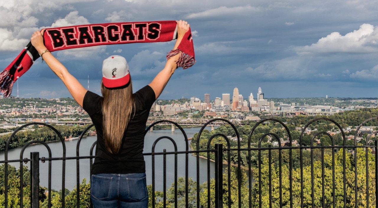 Woman stands facing the city of Cincinnati holding a UC Bearcats scarf in the air.