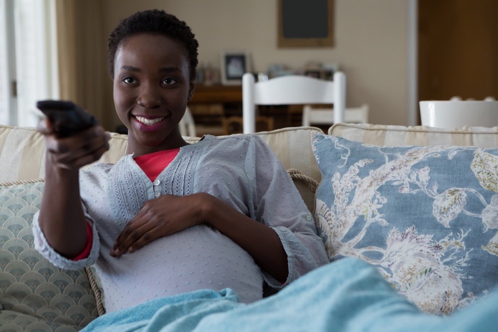 Pregnant woman sits on a couch smiling as she watches TV. photo/Adobestock