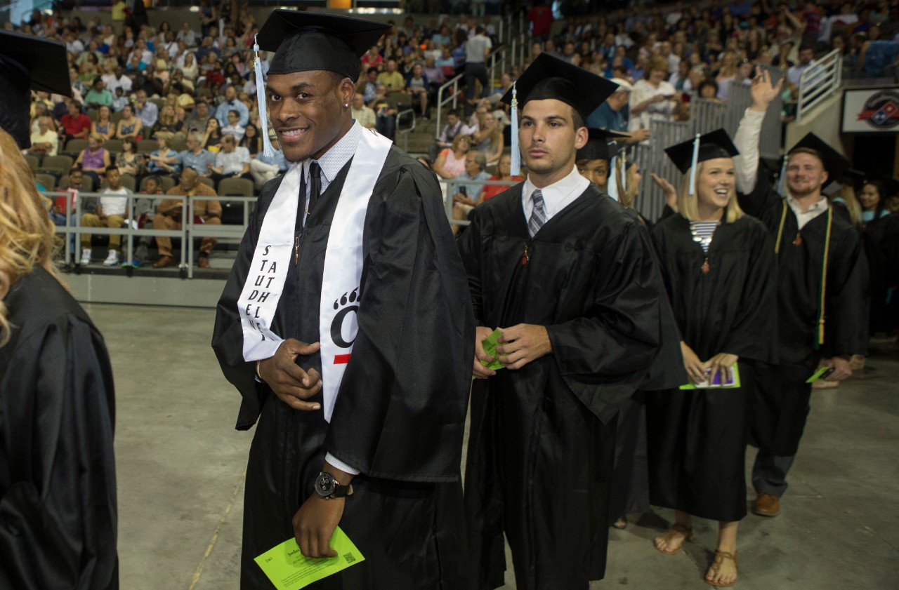 A student athlete graduate smiles and points to the camera among other grads at UC Commencement