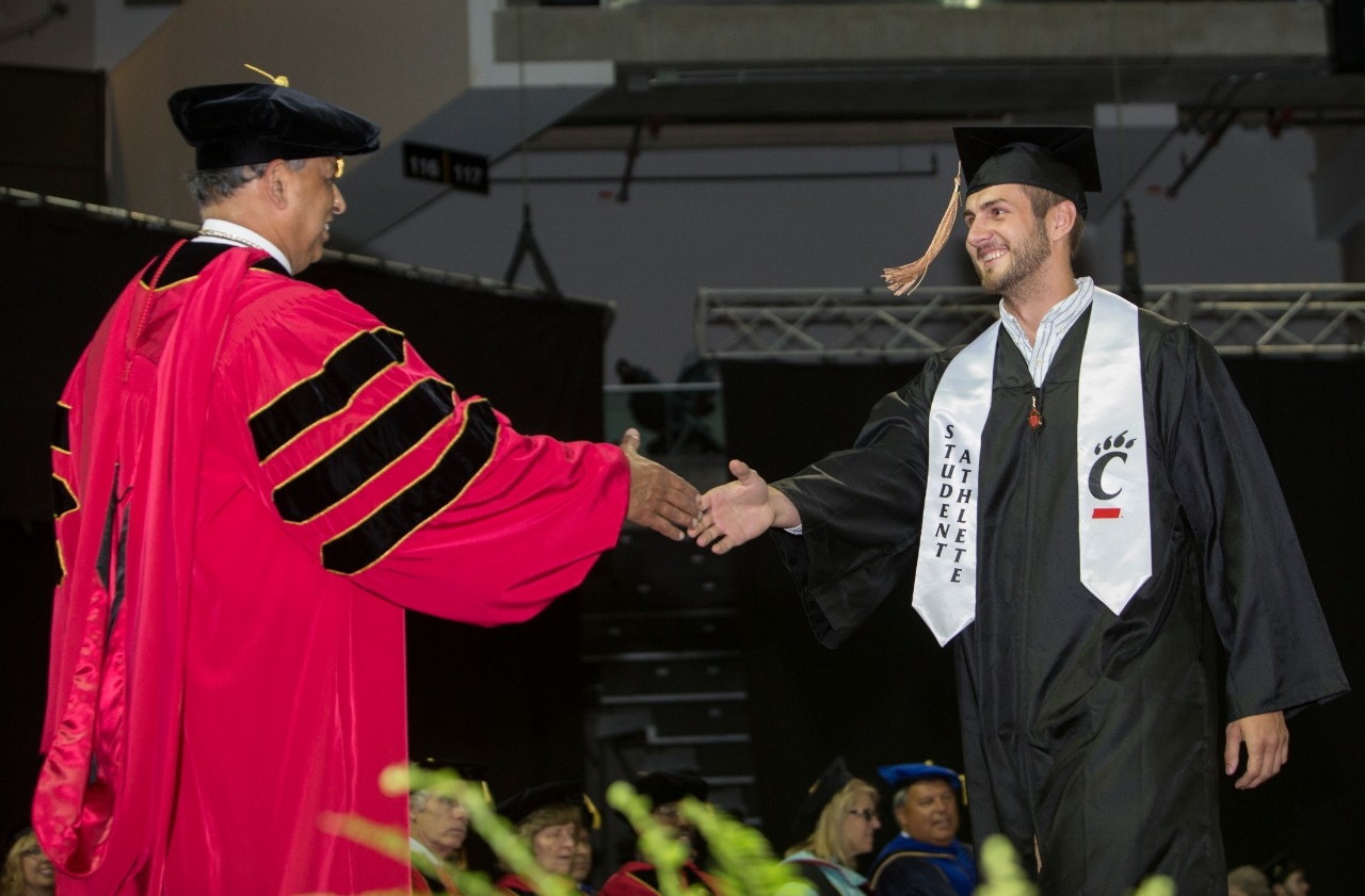 A student athlete graduate shakes president Pinto's hand on stage at UC Commencement.