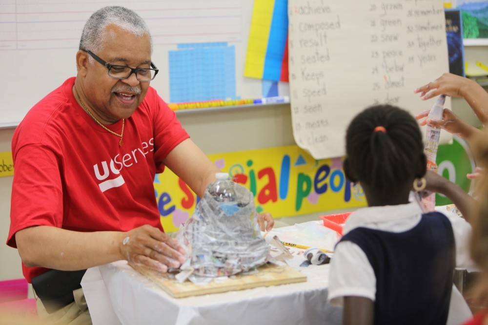 A man sits at a table with a young schoolgirl making volcanoes out of paper and glue.
