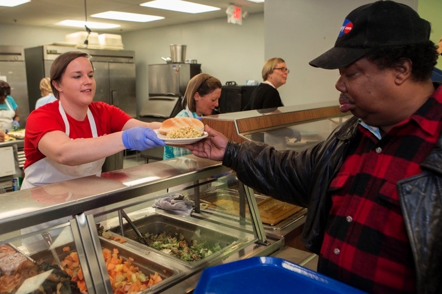 woman hands a man a plate of food across the counter at a local soup kitchen.