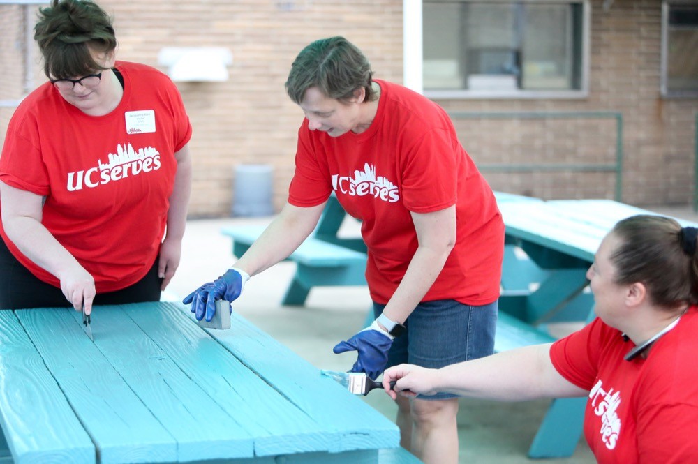 Three UC Serves volunteers paint poolside picnic tables at the Gamble Nippert YMCA on UC Serves Day.