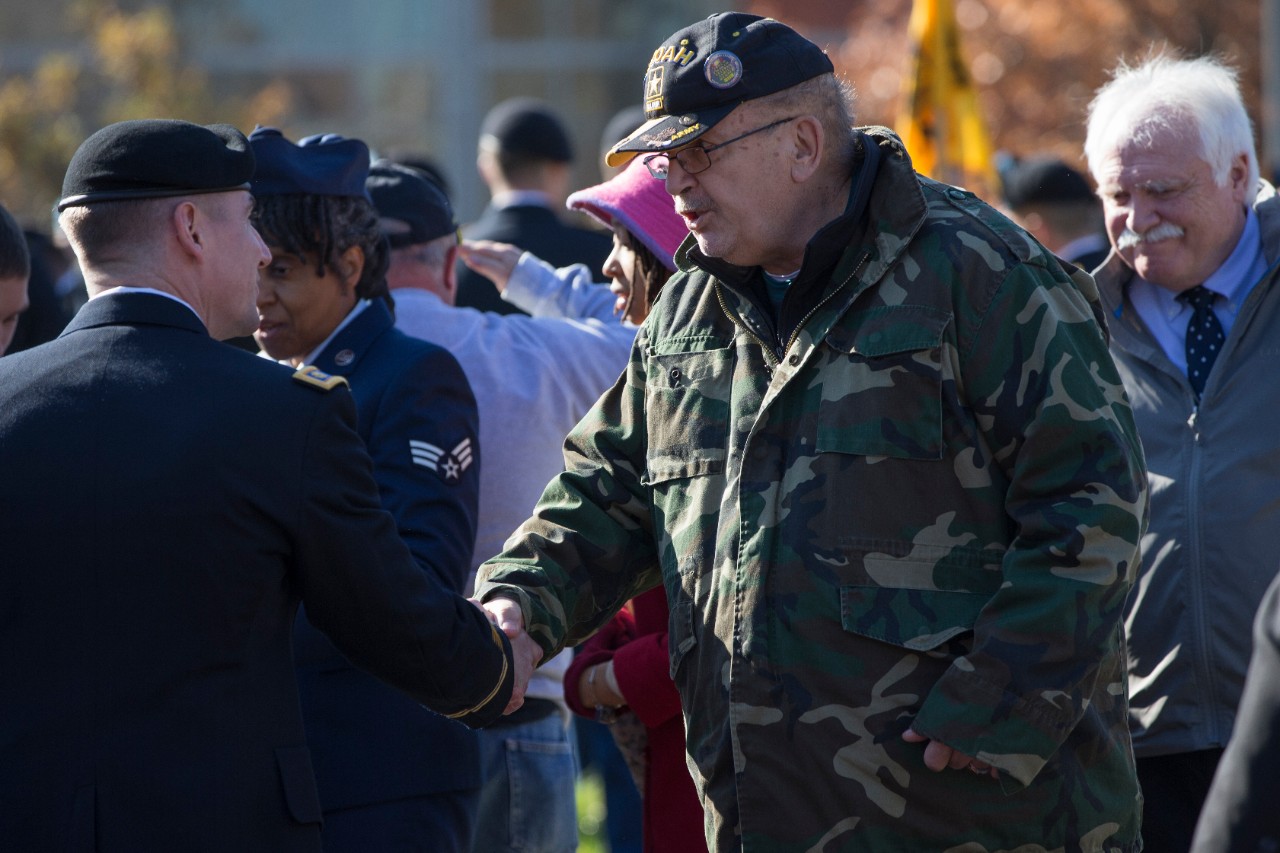 A male uniformed soldier shakes hands with a male attendee wearing a camouflage jacket. 