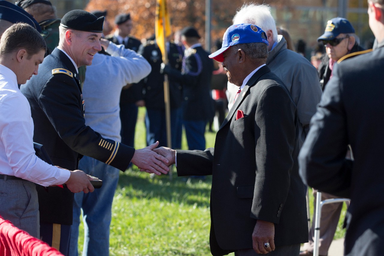 A veteran wearing a blue baseball hat and a suit is given a coin commemorating this year's Veterans Day ceremony. 