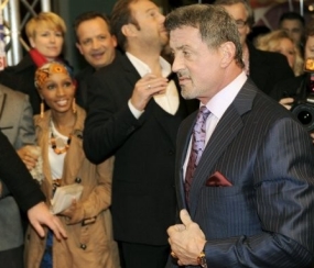 Sylvester Stallone, wearing a suit, walks through a crowd of fans with cameras.