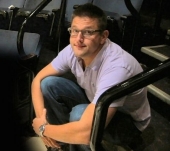 Greg sitting in a theater seat with his legs casually tucked up in the seat and his arms crossed over his knees.