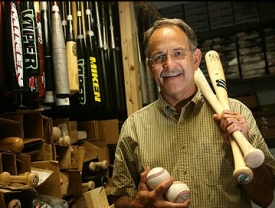 Jm Sherwood holding two baseballs and two bats, with other bats hanging behind him.