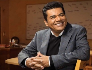 George Lopez is sitting in a student's desk in a classroom.