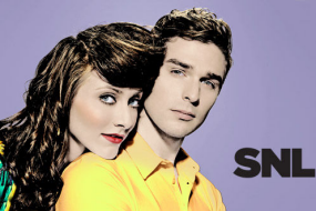 Zooey Deschanel poses for a SNL promo shot leaning on a man and wearing her hair partially piled on top of her head and partially in long curls, too.