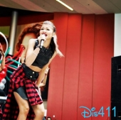 Teenaged pop sgtar sings on stage at an outdoor concert. Her outfit includes a red and black check skirt that resembles a flannel shirt tied around her waist.