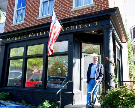 UC alumnus and outstanding architect Mike Watkins stands in front of his brick building next to an American flag.