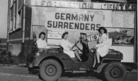 Nurses sitting in a jeep with a large sign behind them that reads: "Germany Surrenders."