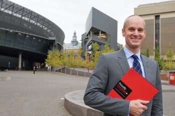 UC Alumnus Drew Becher stands on campus holding a red Proudly Cincinnati folder.