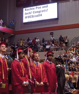 UC graduates in red cap and gowns stand beneath a video board that says, Rachie Poo and Jack! Congrats! #UCGrad14