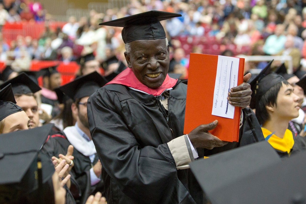 A smiling 82-year-old graduate holds up his copy of his thesis during the University of Cincinnati commencement ceremony.