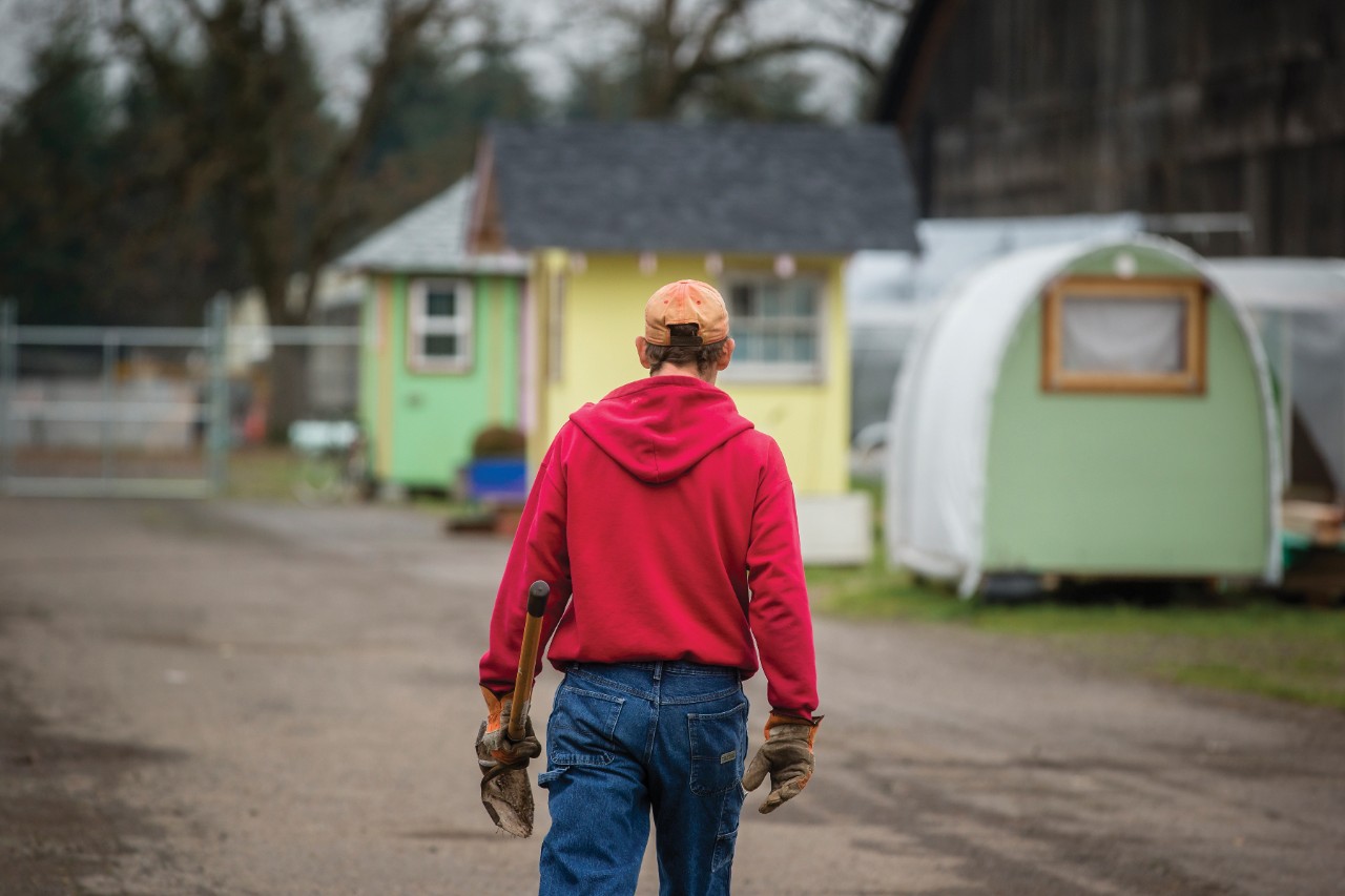 One solitary man in work gloves carrying a shovel walks away from the camera in a new place for homeless called Opportunity Village Eugene (Oregon). 