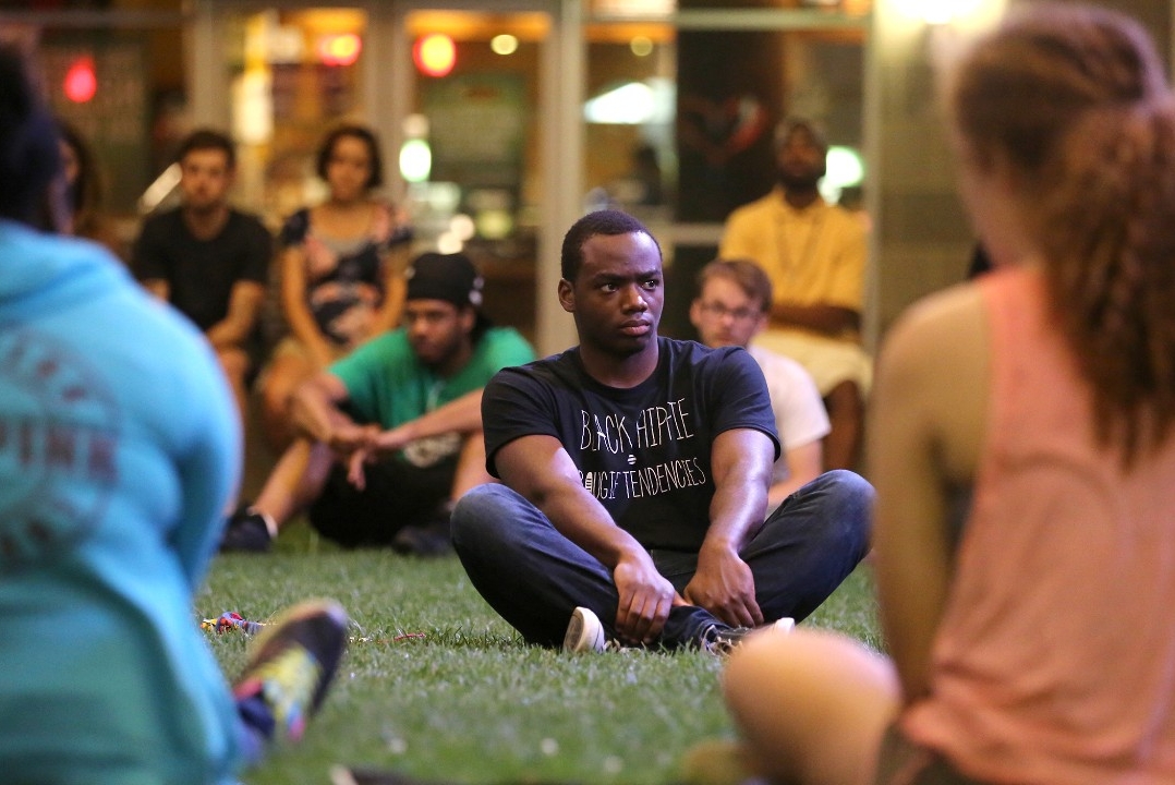 A man sits on the grass in the center of a circle of UC students as part of a peaceful protest at USquare at the Loop near campus.