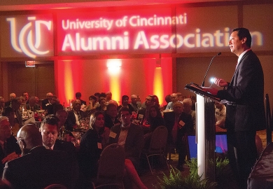 UC President Santa Ono stands at a podium with a dramatic illuminated sign in the background of the UC Alumni Association.