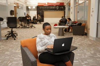 A UC engineering student sits on a cushioned chair looking at her laptop.