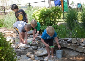 Four boys wade through the brook and dig in the rocks.