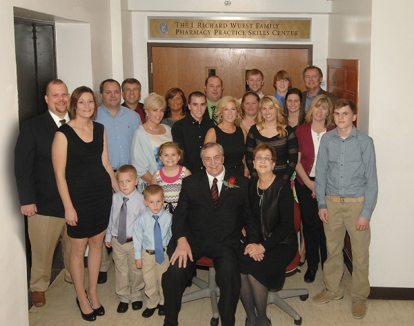 Dick Wuest surrounded by his family members standing in front of the doors of the J. Richard Wuest Family Pharmacy Practice Skills Center at UC.  